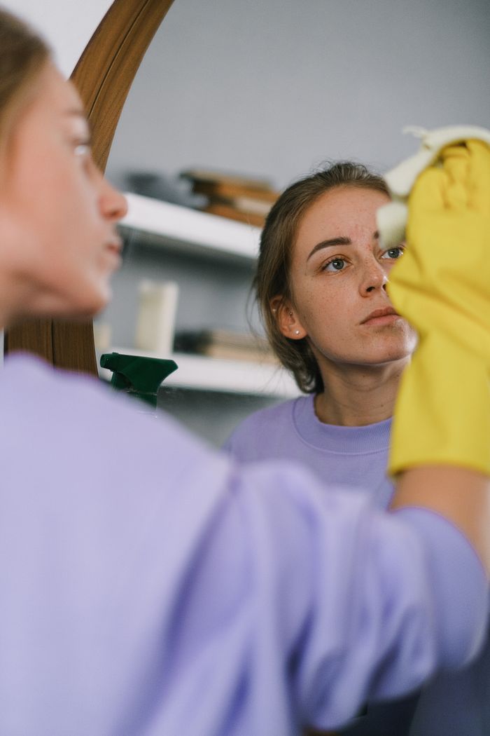 gallery-1 Femme cadrée en plan rapproché, portant un sweat-shirt violet et des gants en latex, nettoyant un miroir dans un appartement lumineux en journée